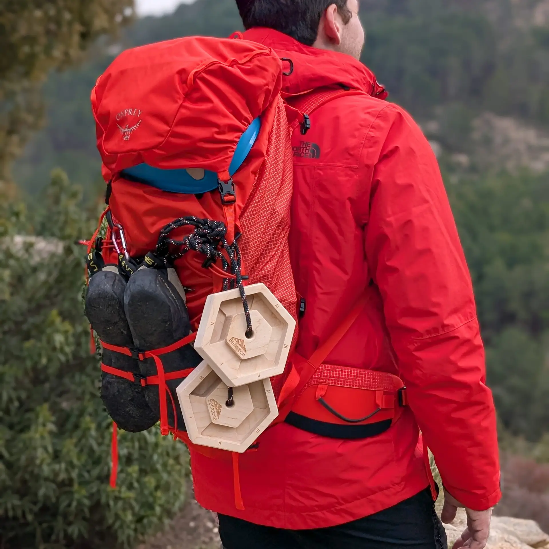 Person wearing a red jacket with two wooden hexagonal hangboards attached to a hiking backpack in a natural outdoor setting.